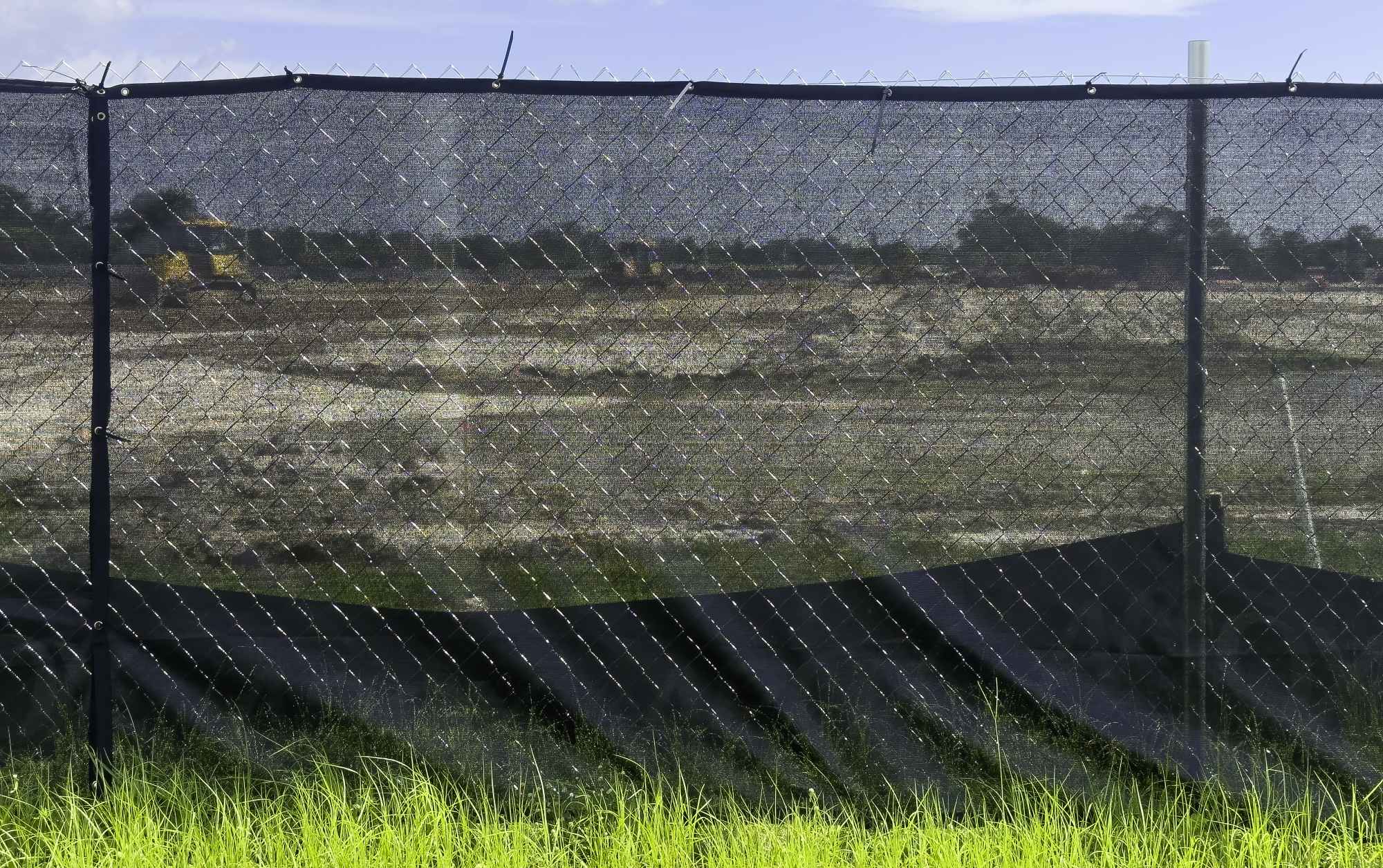 A construction site with dirt and machinery is partially obscured by a black mesh fence; green grass grows in the foreground under a blue sky.