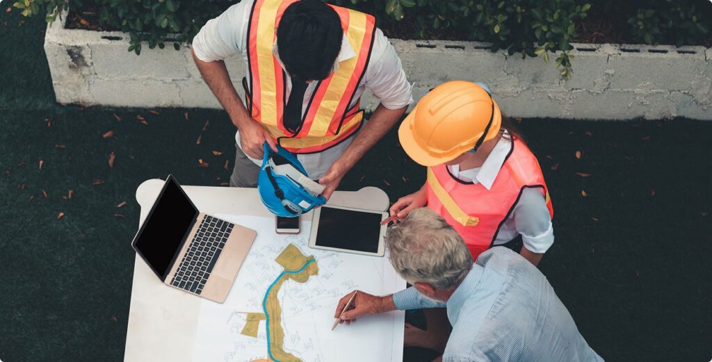 Three people, two in safety vests and one in a hard hat, look at blueprints and maps on a table with a laptop and tablet, appearing to discuss a construction or engineering project outdoors.