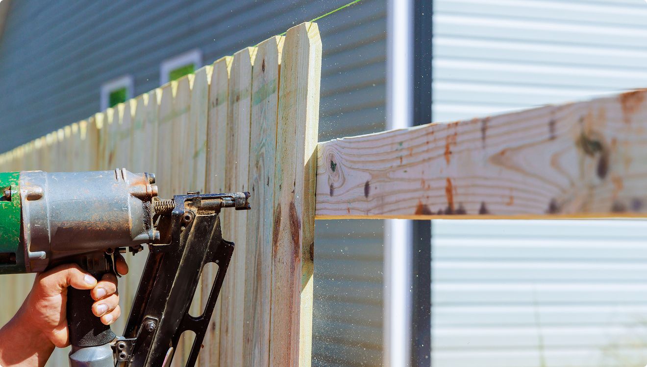 A person uses a nail gun to attach vertical wooden boards to a horizontal support, constructing a wooden fence near a light blue building with horizontal siding.