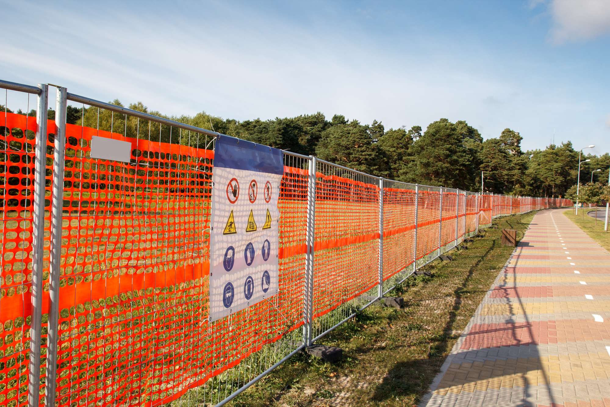 A long orange safety fence with a warning sign blocks off a grassy area next to a paved walkway. Trees and a blue sky with some clouds are visible in the background.