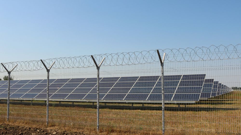 Rows of solar panels are installed in a field behind a tall metal fence topped with barbed wire, under a clear blue sky.