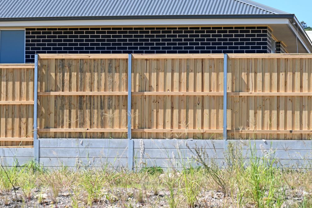 A wooden fence with metal posts stands in front of a modern house with dark brick walls and a gray metal roof. Dry grass and weeds grow in the foreground.