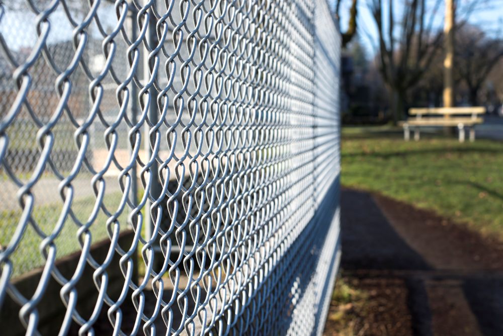Close-up of a chain-link fence with a park in the background, showing a pathway, green grass, and a bench under trees on a sunny day.