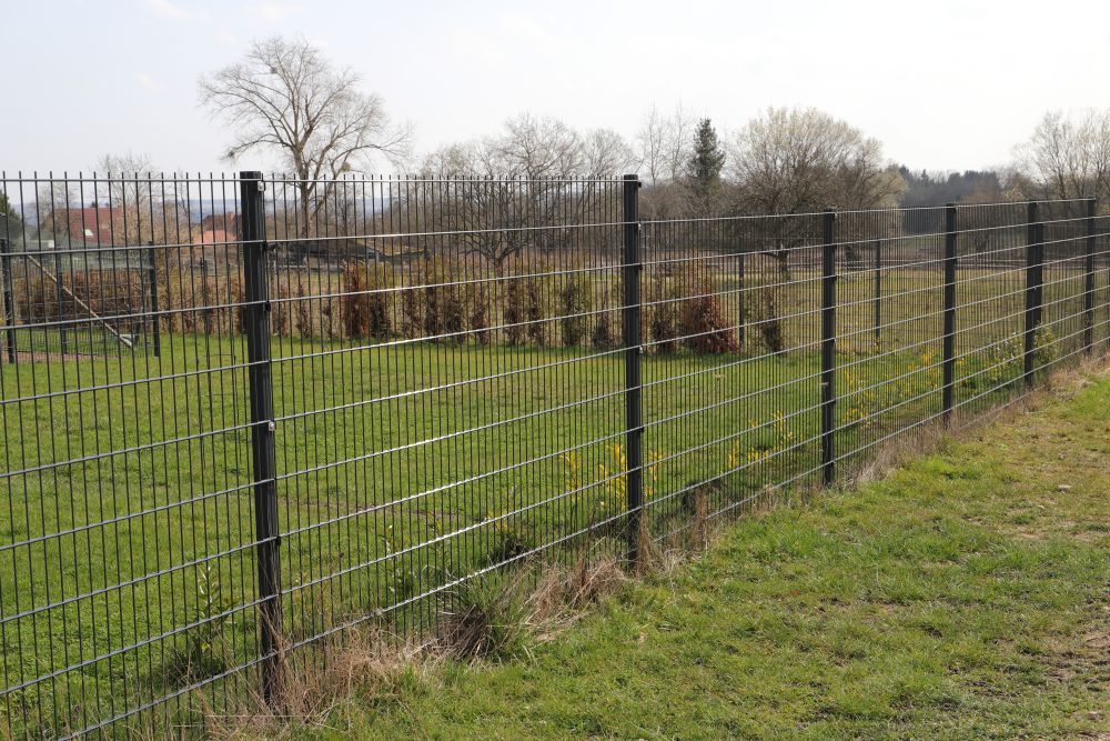 A metal wire fence runs along a grassy area with trees and bushes in the background on a partly cloudy day.
